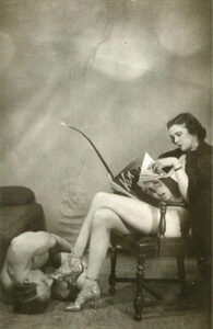 Fotografia de Charles Guyette. Uma mulhere está sentada lendo uma revista e segurando um chicote enquanto um homem está no chão beijando os pés dela.